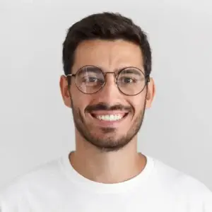 Daylight portrait of young handsome caucasian man isolated on grey background, dressed in white t-shirt and round eyeglasses, looking at camera and smiling positively